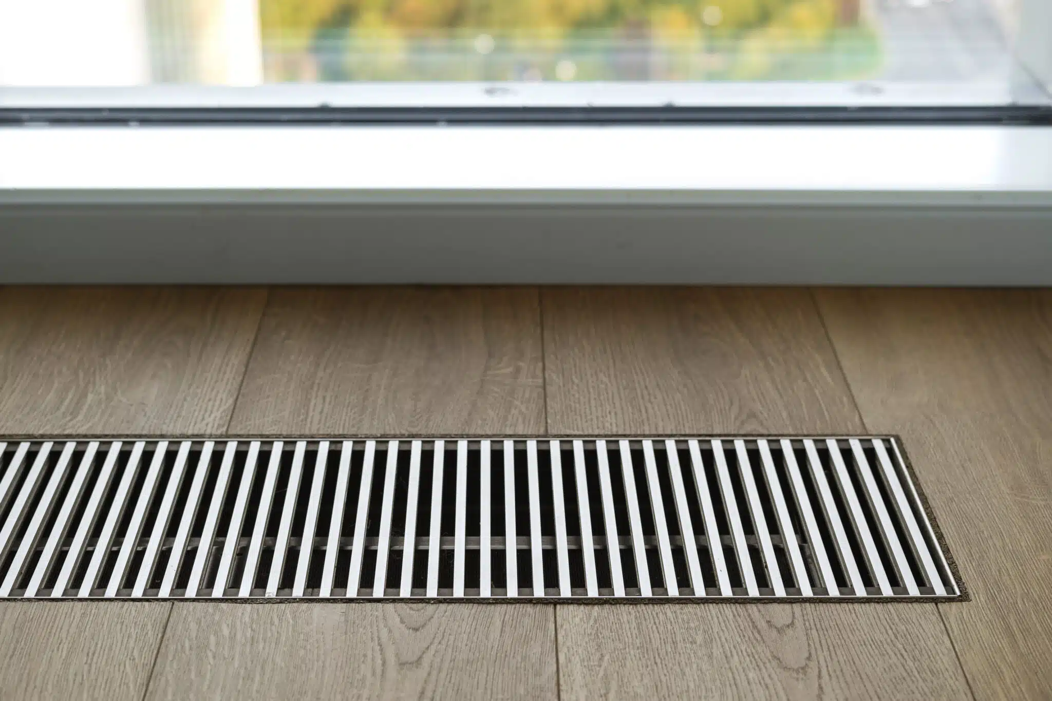 Close up of wooden floor with chrome ventilation grid of central heating against window.